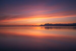 A vibrant sunset casts orange and purple hues over a calm, reflective body of water with distant mountains on the horizon.