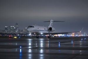 A private jet taxis on a wet airport runway at night with a city skyline in the background.