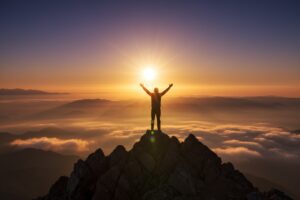 A person stands on a mountain peak with arms raised toward the rising sun above a sea of clouds.